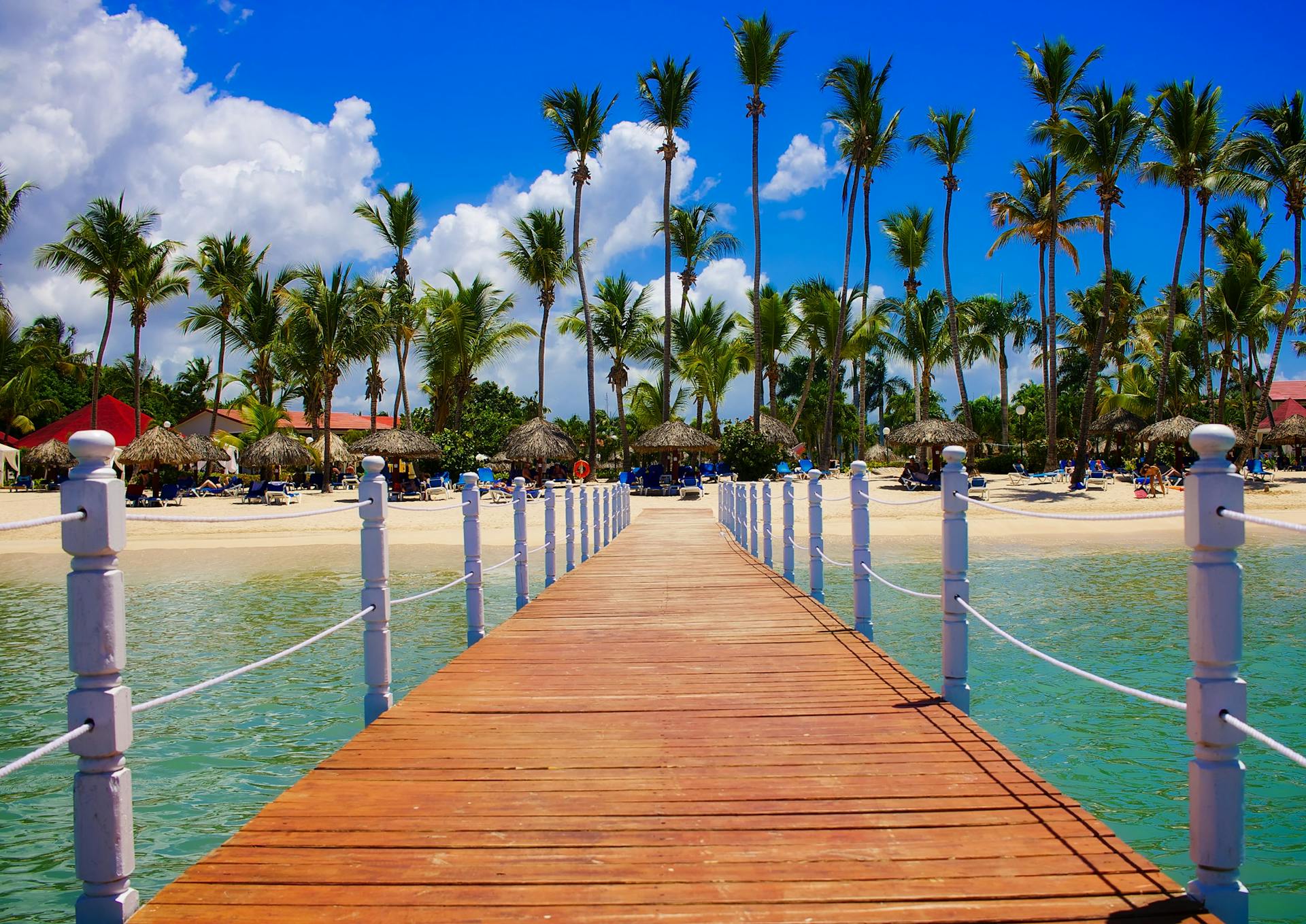 Turismo República Dominicana 2025: pasarela de madera hacia playa de aguas turquesas en Punta Cana, con palmeras y cielo azul.